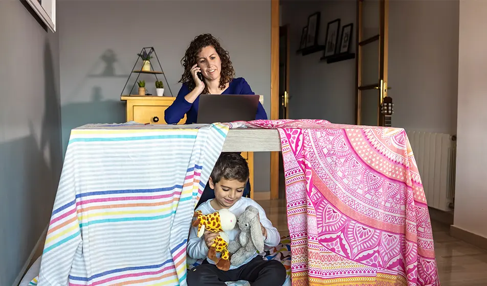 A woman is working from home and is sat at her desk with her laptop. In front of her, her child has covered a desk with colourful sheets and has made it into a den. The child is playing with a giraffe and listening to his mother working.